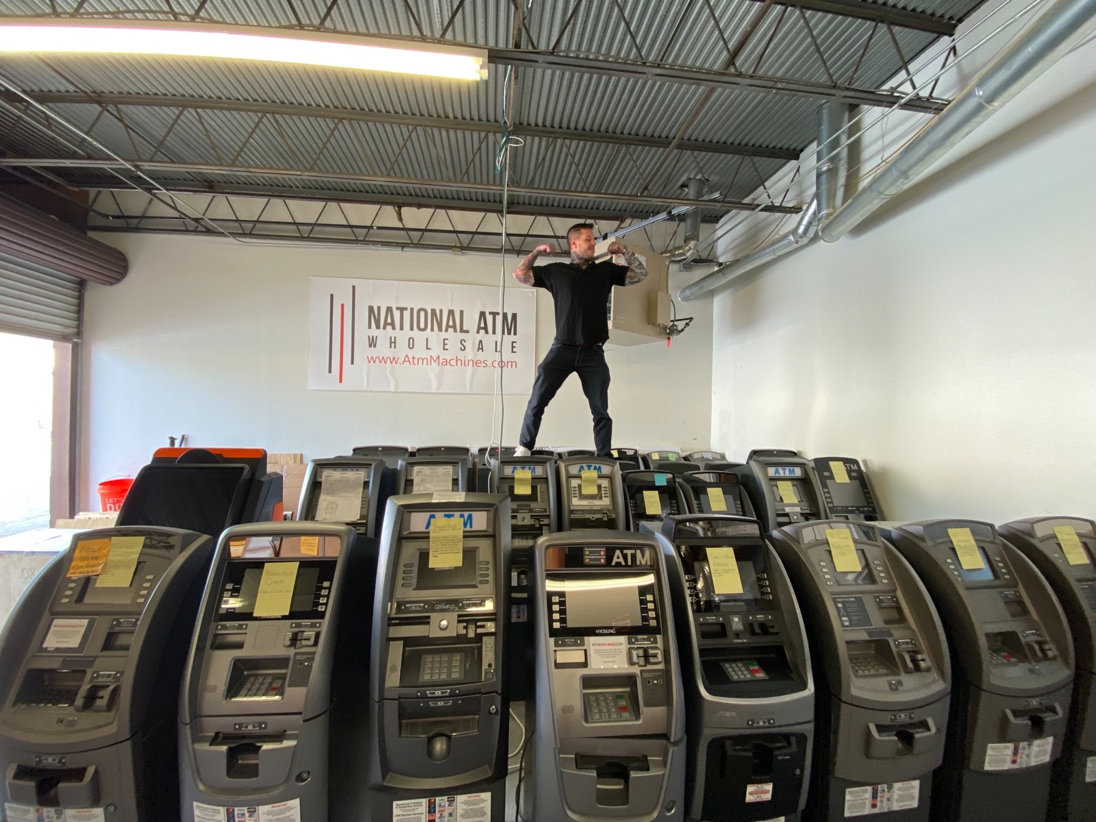 Justin Gilmore standing on top of rows of ATMs in a warehouse, beneath a banner reading www.AtmMachines.com